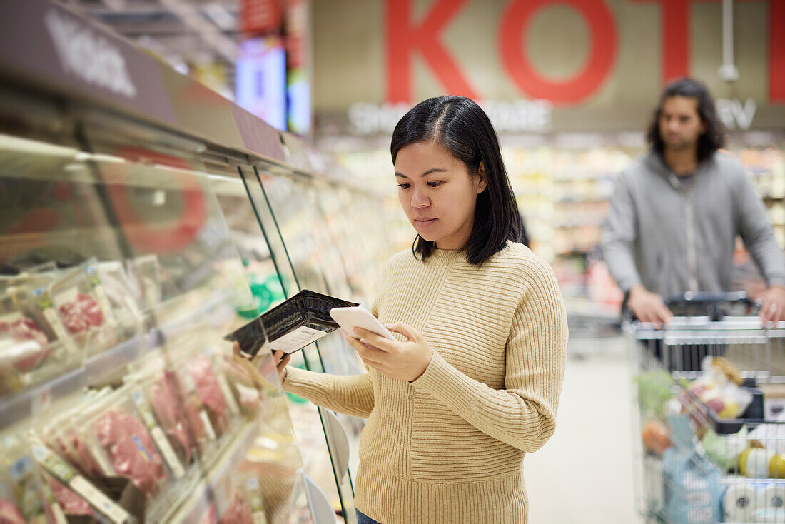 Frau kauft im Supermarkt ein und benutzt ihr Handy, um Preise zu vergleichen oder die Einkaufsliste zu prüfen