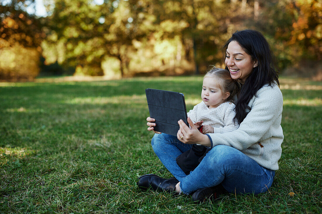 Mother and daughter sitting in autumn park and using digital tablet\n