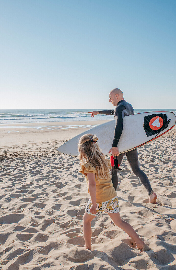 Blond girl with father carrying surfboard on beach\n