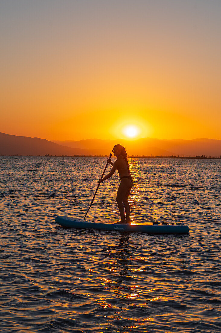 Silhouette einer jungen Frau, die bei Sonnenuntergang am Strand von Trabucador, Ebro-Delta, Tarragona, Spanien, Paddelsurfen übt