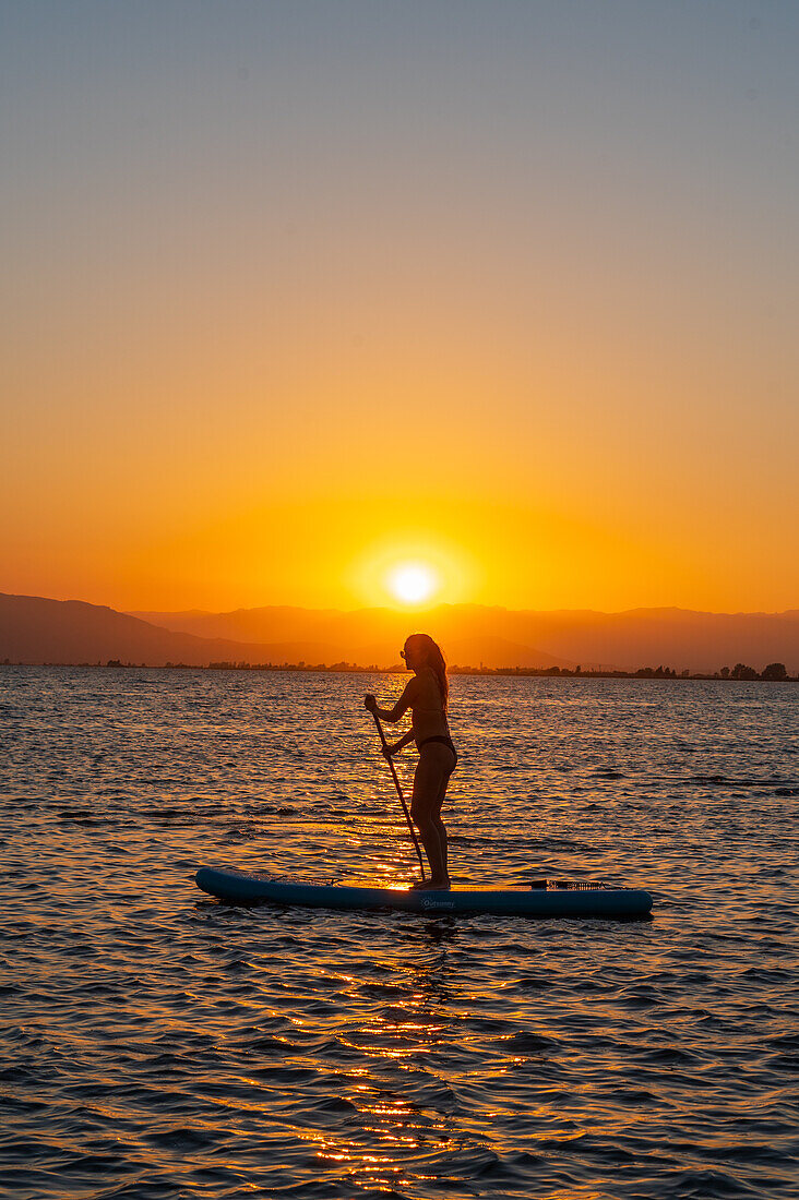 Silhouette einer jungen Frau beim Paddelsurfen während des Sonnenuntergangs am Strand von Trabucador, Ebro-Delta, Tarragona, Spanien