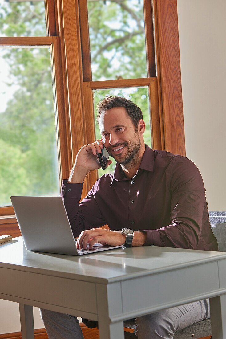 Smiling man using laptop and smart phone at table at home\n