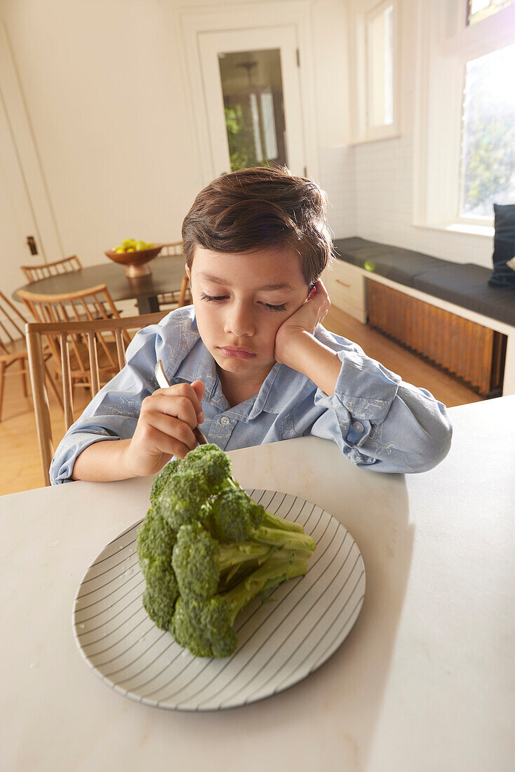 Unzufriedener Junge (8-9) betrachtet Brokkoli auf einem Teller in der Küche