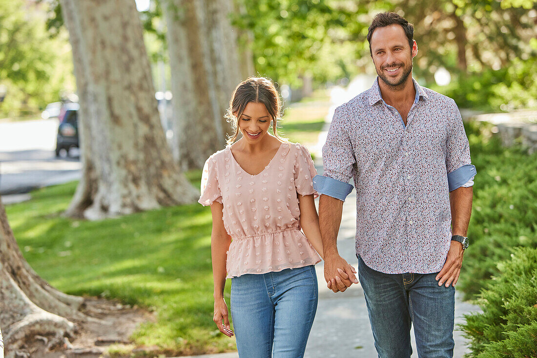 Smiling couple holding hands, walking on treelined sidewalk\n