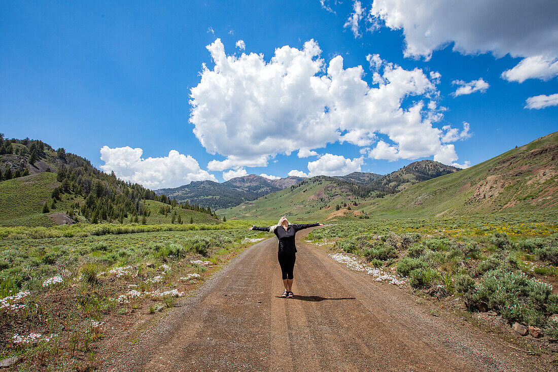 USA, Idaho, Sun Valley, Senior woman standing with arms outstretched on road in mountain scenery\n