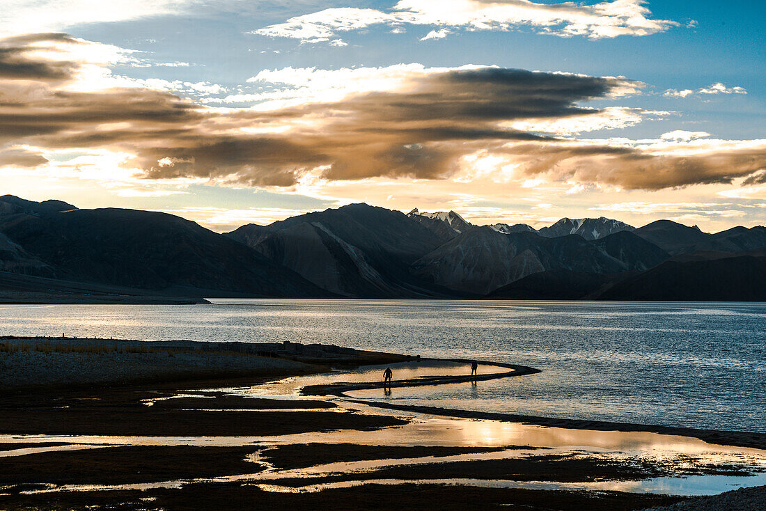 Sunset at Pan Gong lake with golden light reflection in the flat waters showing two people walking along the shore, Ladakh, northern India, Asia\n
