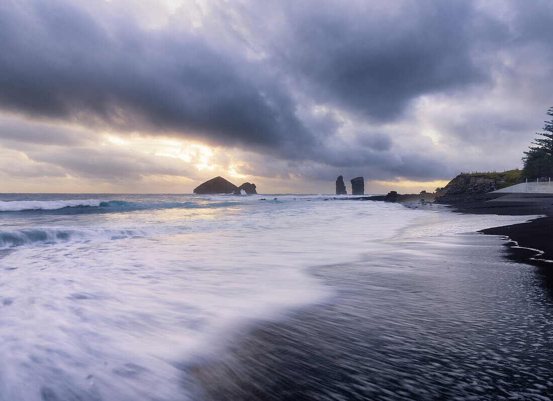 Langzeitbelichtung der Wellen am schwarzen Sandstrand von Mosteiros bei bewölktem Sonnenuntergang mit den Meeresstapeln im Hintergrund, Mosteiros, Sao-Miguel-Insel, Azoren-Inseln, Portugal, Atlantik, Europa