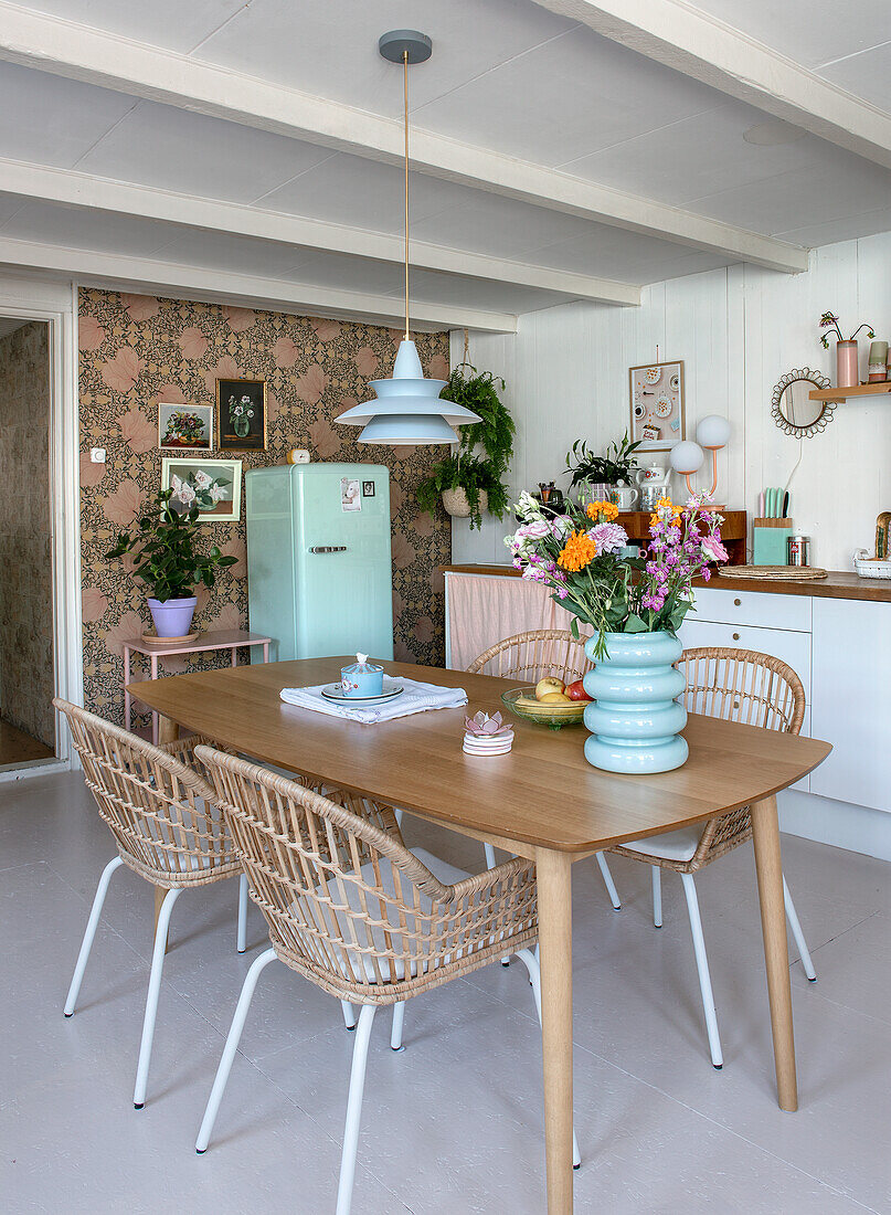 Dining area with wooden table, rattan chairs and bouquet of flowers in kitchen with retro-style patterned wallpaper