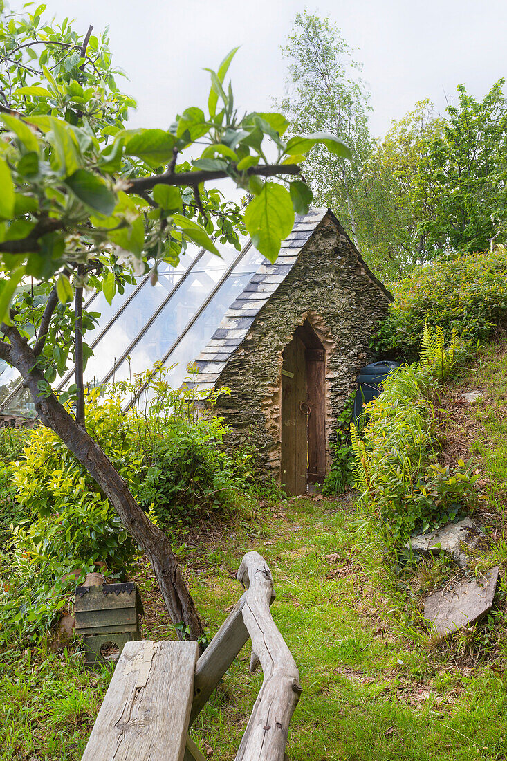 Gartenhaus aus Stein mit Glasdach neben Obstbaum und Holzbank im Garten