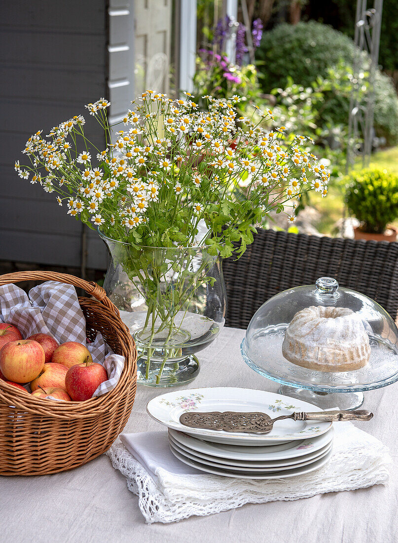 Laid garden table with apple basket, bouquet of daisies and cake