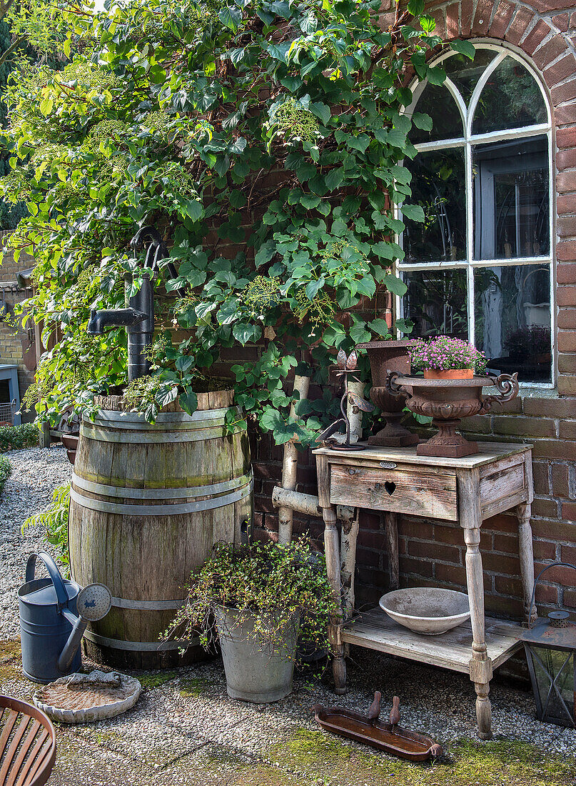 Rustikaler Gartentisch mit Blumenkübel und alter Weinfasspumpe vor Backsteinfassade