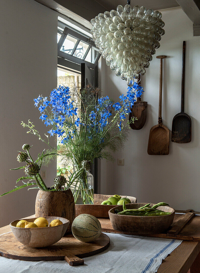 Kitchen table with bowls full of fruit, wooden utensils and blue delphinium