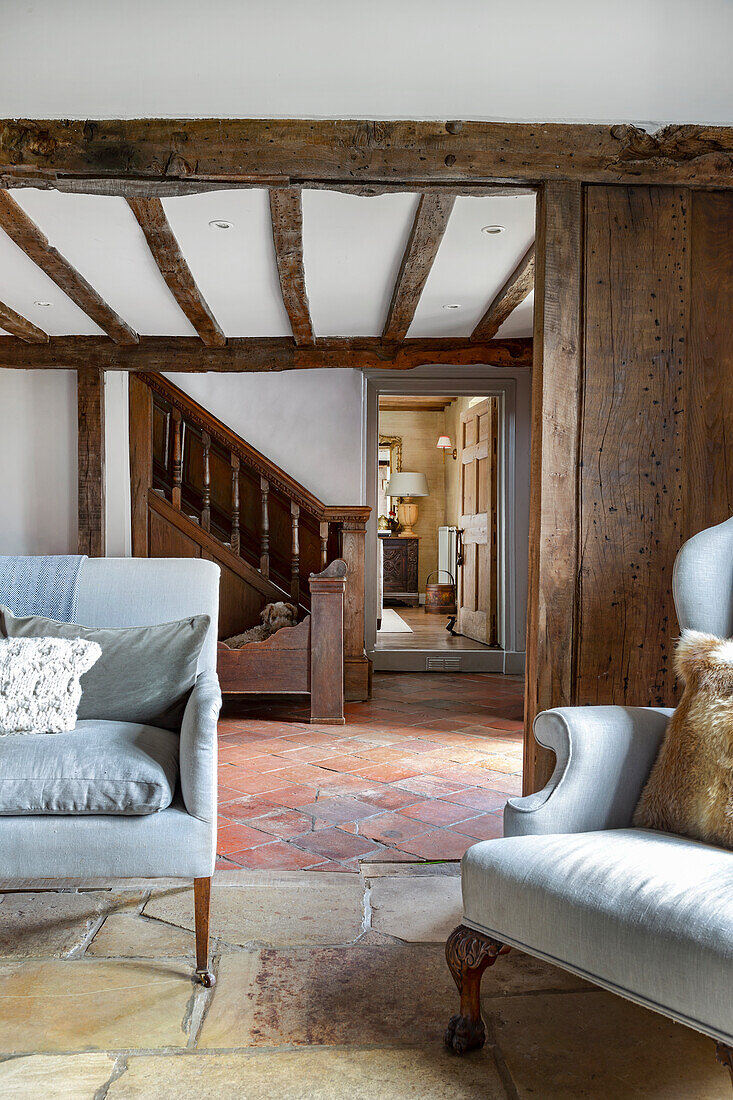 Living area with light-colored upholstered furniture and exposed wooden beams, view of hallway with terracotta tiles and wooden staircase