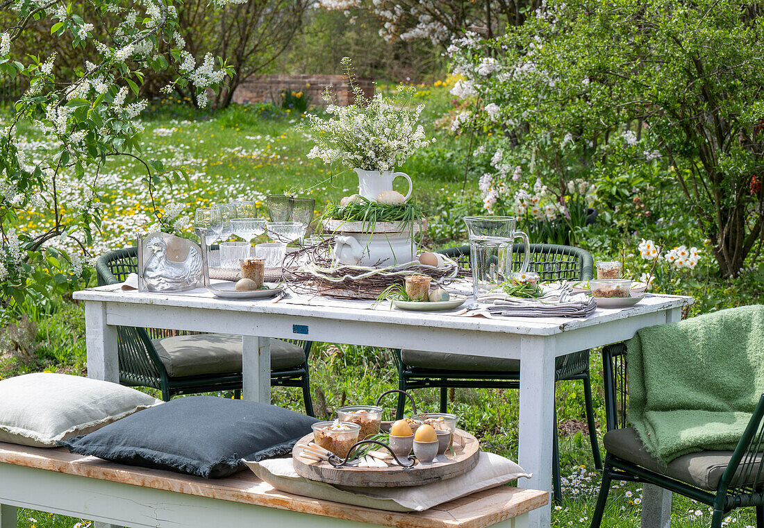 Gedeckter Tisch im Garten für Osterfrühstück mit Osternest und gefärbten Eiern in Eierbechern und Blumenstrauß in Etagere