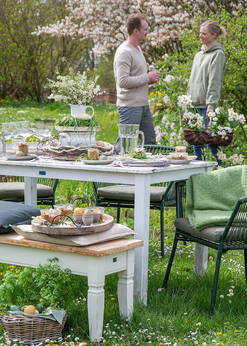 Young couple behind table laid for Easter breakfast with Easter nest and colored eggs in egg cups, daffodils and parsley in a basket in the garden