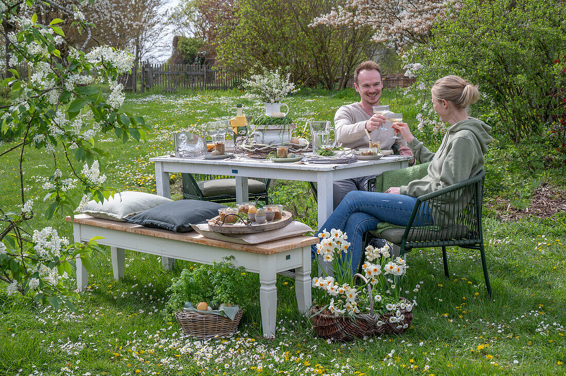 Junges Paar sitzt an gedecktem Tisch zum Osterfrühstück mit Osternest und gefärbten Eiern in Eierbechern, mit Sekt anstoßen, Narzissen und Petersilie im Korb  im Garten