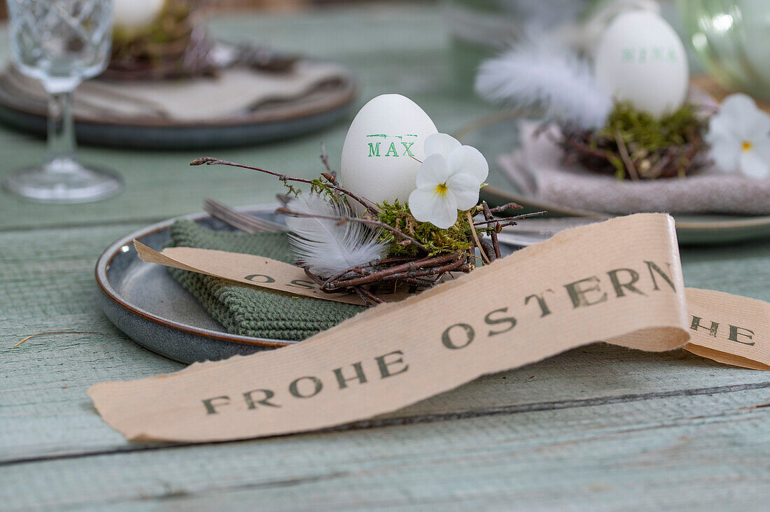 Easter egg with name on small nest with feathers and violet flowers, on plate with napkin and Easter greeting