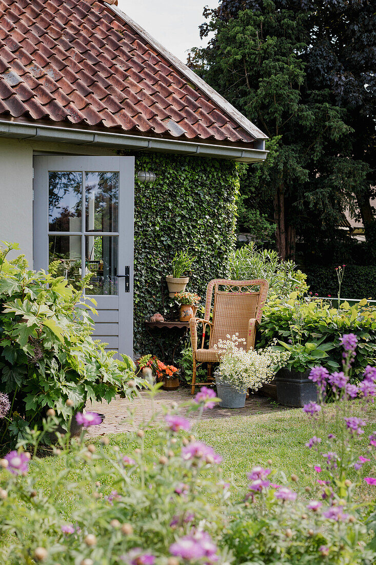 Kleiner Sitzbereich mit Rattan-Sessel und Blumentöpfen im Garten vor einem begrünten Haus