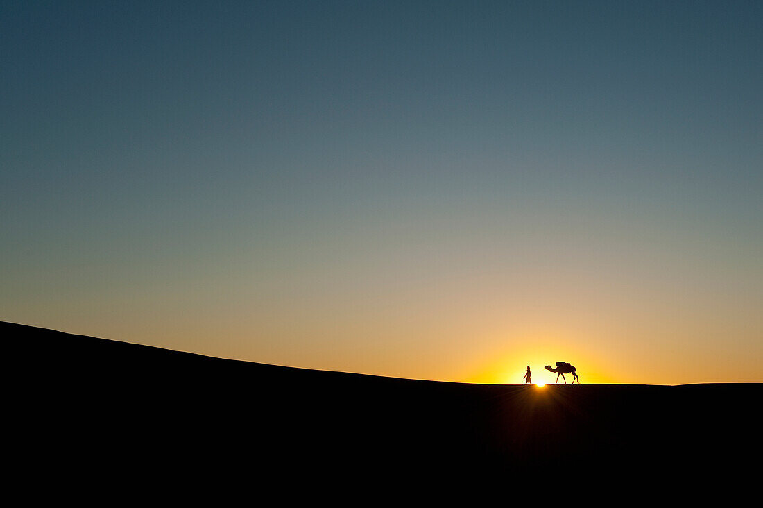 Marokko,Silhouette von Berber "Blauer Mann" führt Kamel über Sanddünen in der Abenddämmerung in Erg Chebbi Bereich, Sahara-Wüste in der Nähe von Merzouga