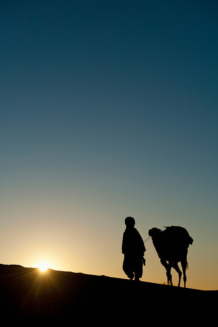 Morocco,Silhouette of Berber 'Blue man' leading camel across sand dunes at dusk in Erg Chebbi area,Sahara Desert near Merzouga