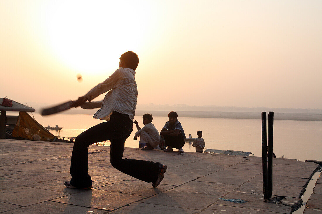 Einheimische Jungen spielen bei Sonnenaufgang den beliebten Volkssport Cricket am Badeghat. Ein guter Schlag und der Ball fließt in den Ganges. Die Kultur von Varanasi ist eng mit dem Fluss Ganges und seiner religiösen Bedeutung verbunden.