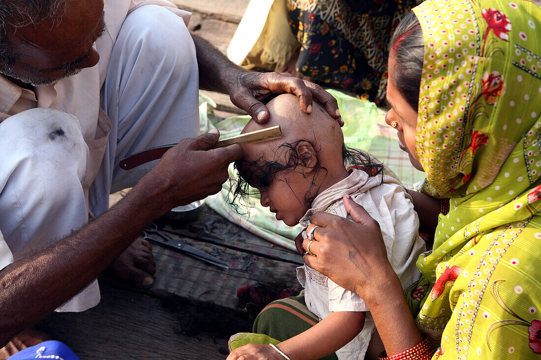 Ein Junge lässt sich von einem Straßenfriseur am Badeghat oberhalb des Ganges den Kopf rasieren. Hindu-Tradition, sich zu günstigen Zeiten den Kopf rasieren zu lassen. Die Kultur von Varanasi ist eng mit dem Fluss Ganges und seiner religiösen Bedeutung verbunden.