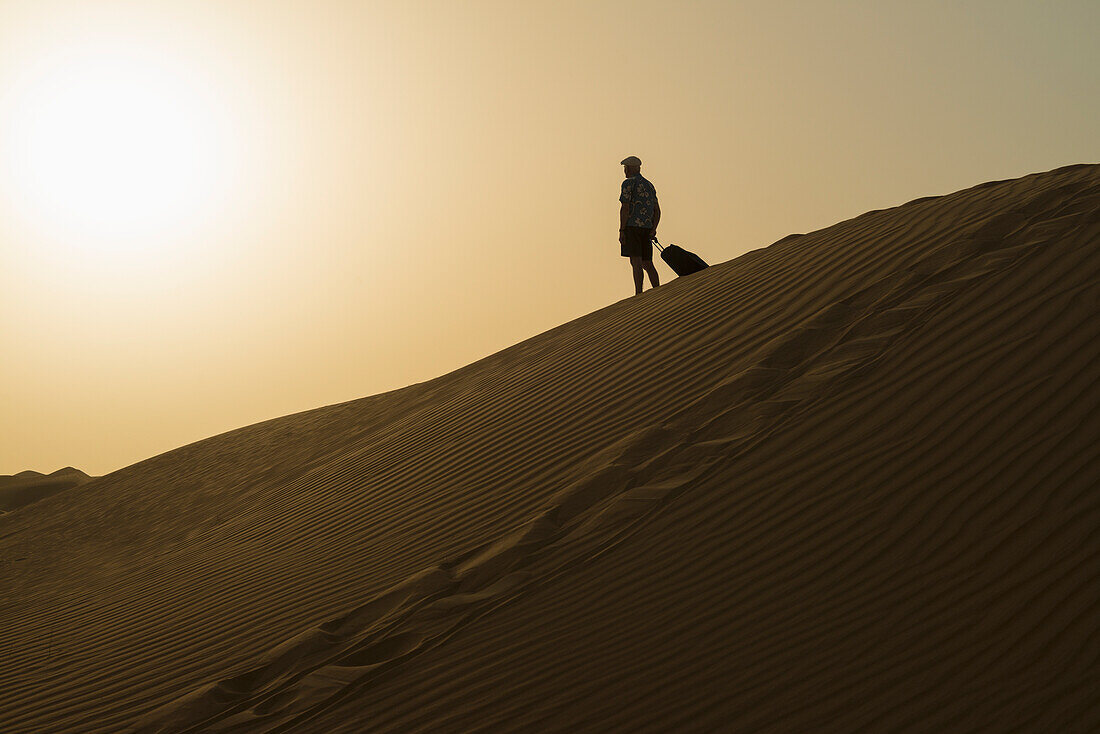Barfüßiger Mann mit Koffer auf Sanddüne in der Abenddämmerung,Dubai,Vereinigte Arabische Emirate