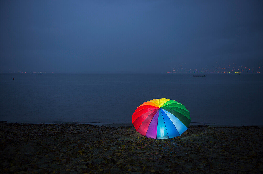 Ein bunter Regenbogen-Regenschirm leuchtet nachts am Ufer des Lago Maggiore, Locarno, Tessin, Schweiz