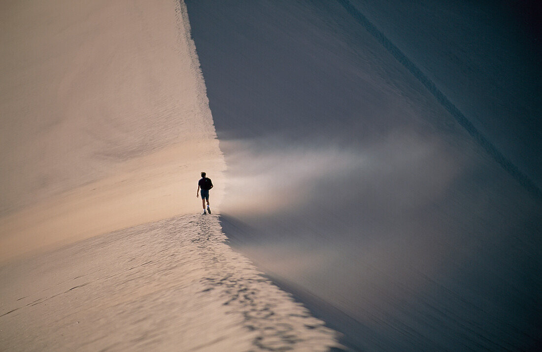 A figure walks into the path of blowing sand on the crest of a dune,Valley of the Moon,Atacama Desert,Chile