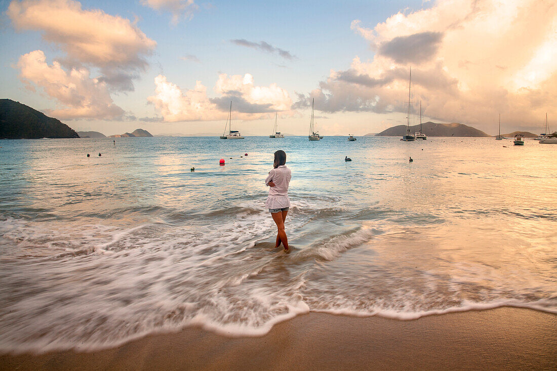 Blick von hinten auf eine Frau, die in der schäumenden Brandung am Strand steht und auf das türkisfarbene Wasser mit Booten schaut, die vor dem Ufer entlang des Horizonts in der Cane Garden Bay in der Abenddämmerung vertäut sind, Tortola, Britische Jungferninseln, Karibik