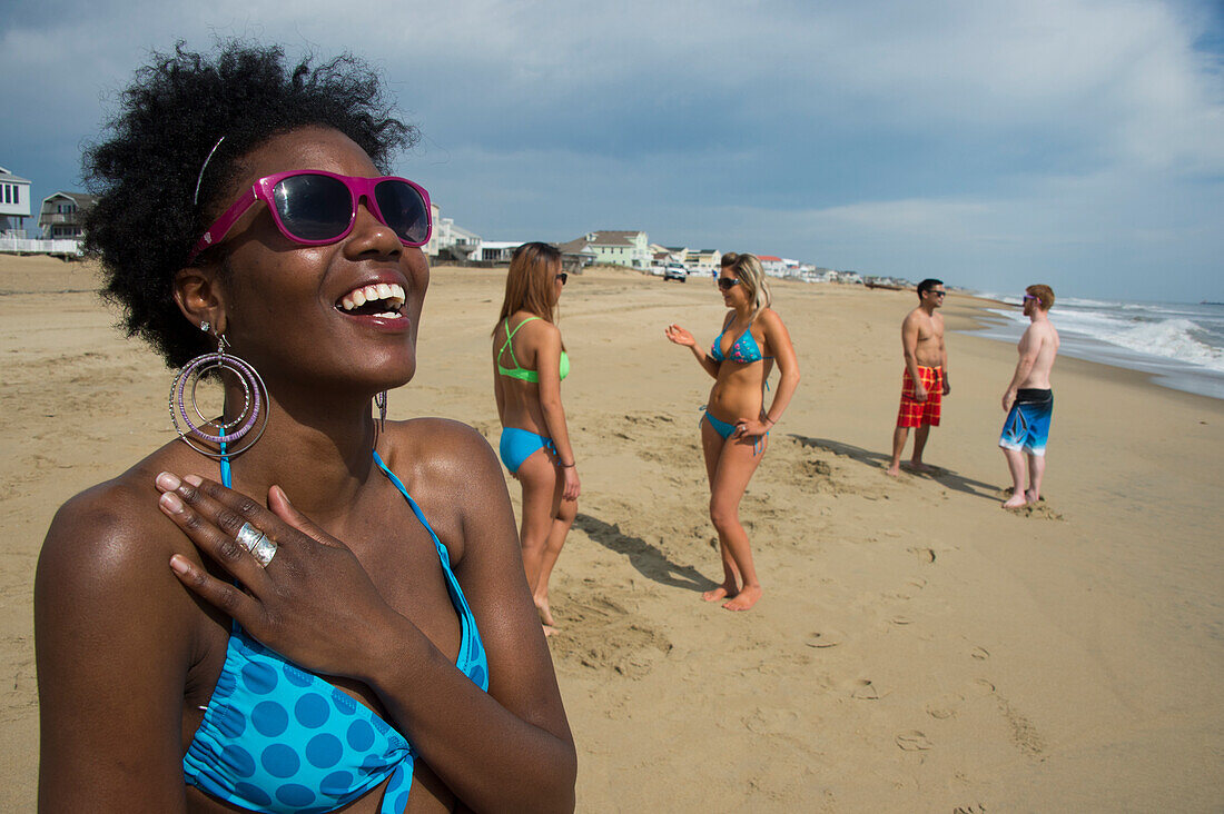 Gruppe von Freunden in Badekleidung am Virginia Beach, mit einer jungen Frau im Vordergrund, die lächelt und eine rosa gerahmte Sonnenbrille trägt, First Landing State Park, Virginia, USA, Virginia Beach, Virginia, Vereinigte Staaten von Amerika