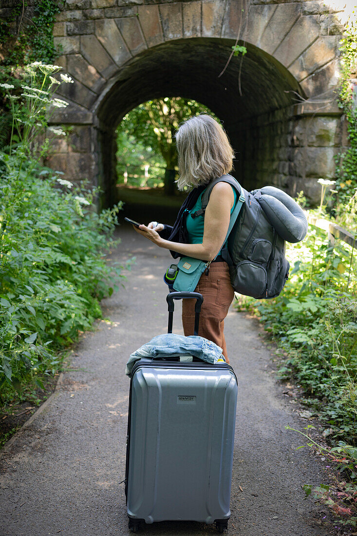 Mature woman uses her smart phone while standing on a path with her luggage,United Kingdom
