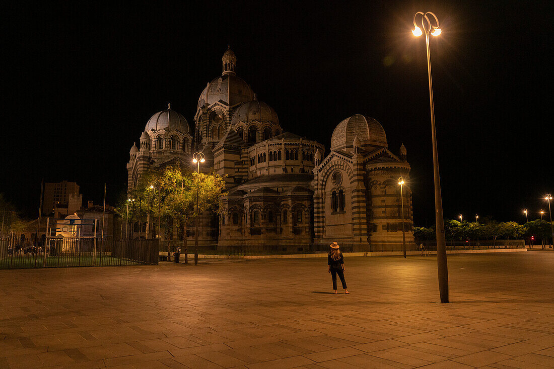 Frau steht und bewundert die Kathedrale von Marseille bei Nacht, mit einer Straßenlaterne, die hell in der Dunkelheit leuchtet, Marseille, Frankreich