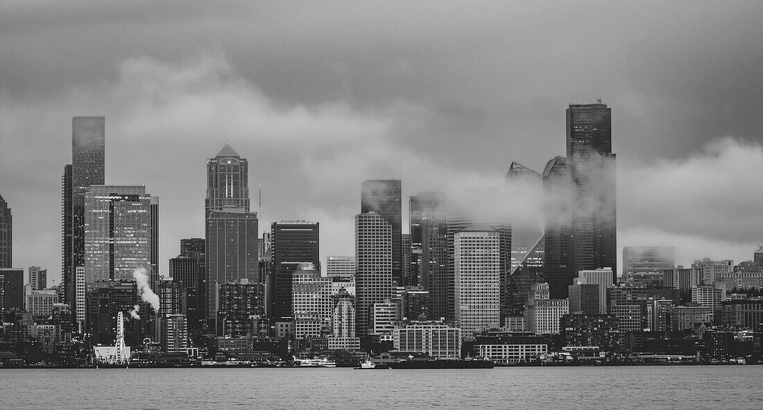 Skyline von West Seattle über die Elliott Bay an einem regnerischen Nachmittag mit niedrigen Wolken und Nebelschwaden, die durch die hohen Gebäude driften, Seattle, Washington, Vereinigte Staaten von Amerika