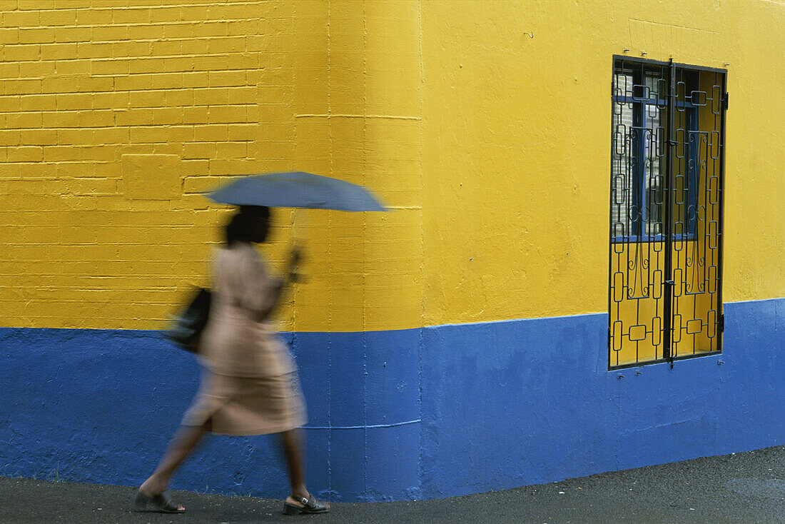 Frau läuft an einer gelben und blauen Wand vorbei und hält einen Regenschirm an einem regnerischen Tag, Grenada, Westindien