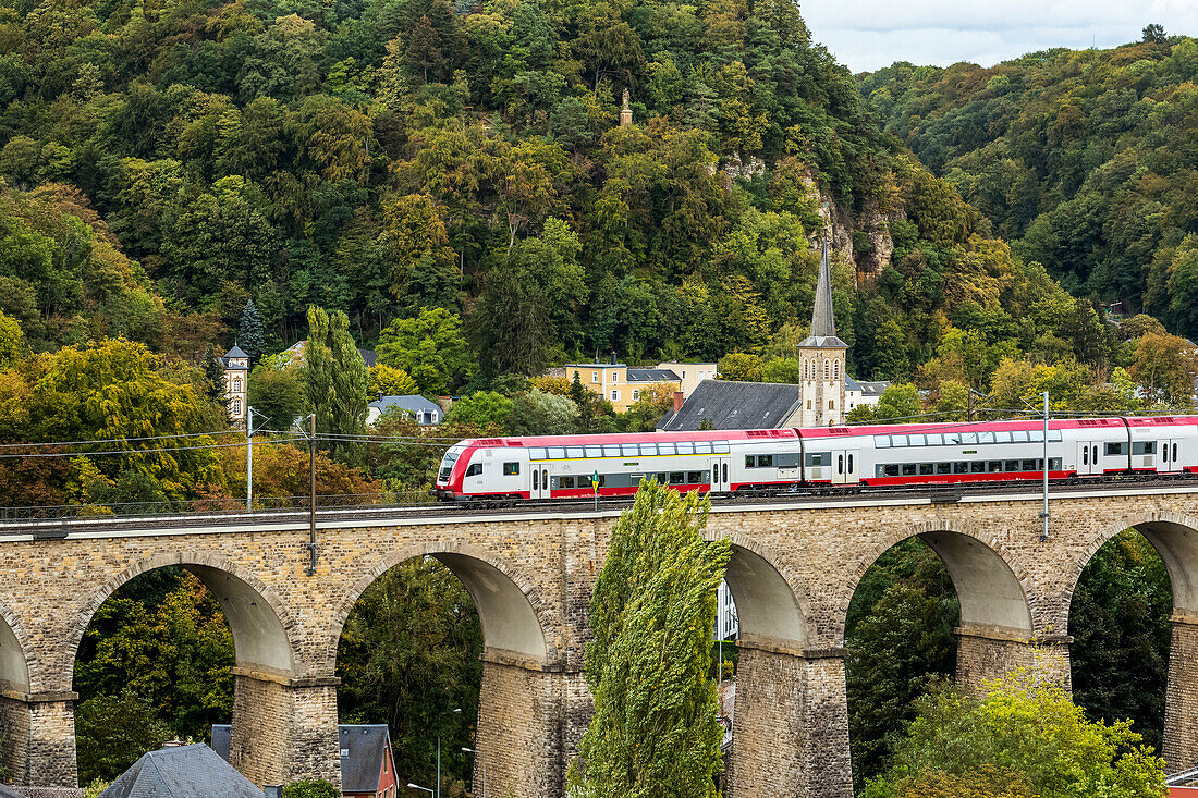 Electric train on old stone arch bridge with church steeple and treed hillside in the background,Luxembourg City,Luxembourg