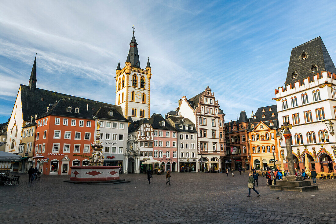 Spaziergänger auf dem mittelalterlichen Marktplatz in einem Dorf mit dekorativem Brunnen, bunten Gebäuden und einem großen Uhrenturm mit blauem Himmel und Wolken, Trier, Deutschland