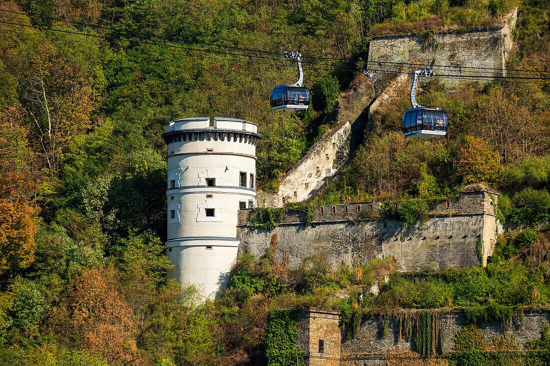 White circular tower on treed hillside with old stone wall and gondolas along cables,Koblenz,Germany