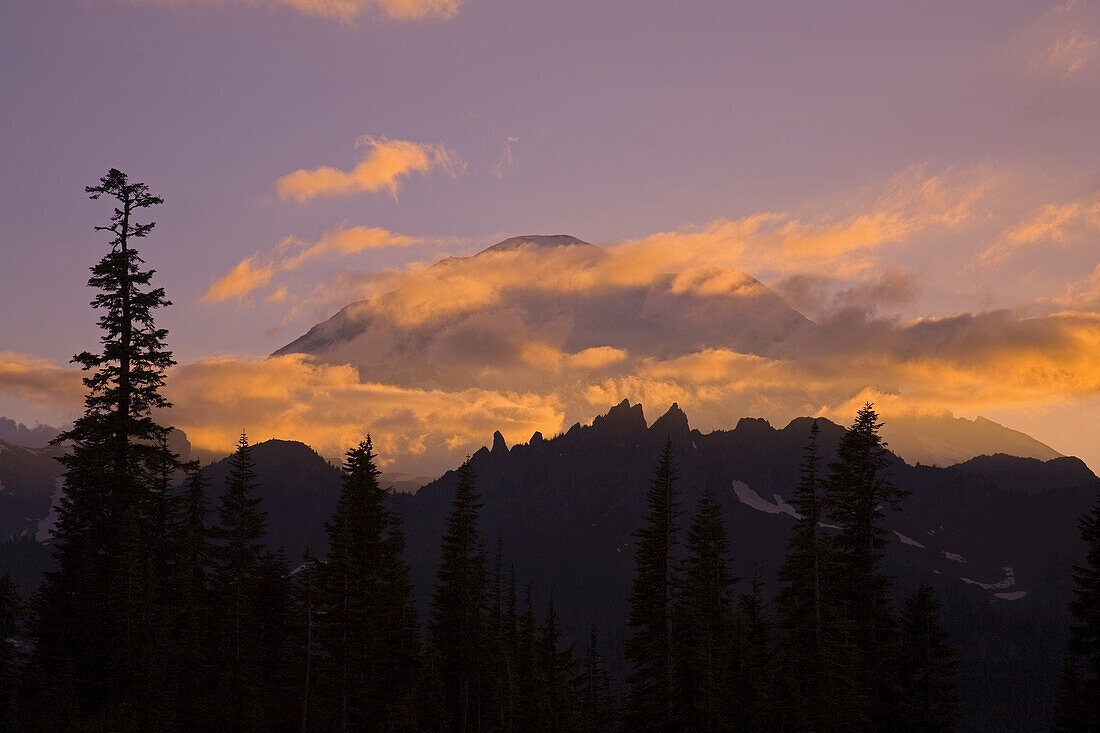 Mount Rainier verdeckt von glühenden Wolken bei Sonnenuntergang, mit Bergen der Cascade Range und Bäumen im Vordergrund, Mount Rainier National Park, Washington, Vereinigte Staaten von Amerika