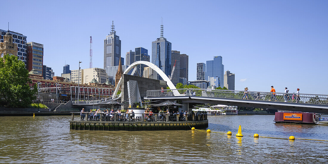 Skyline of buildings in Melbourne viewed from the Yarra River waterfront with a restaurant called Ponyfish Island on the water beside the Evan Walker pedestrian bridge,Melbourne,Victoria,Australia