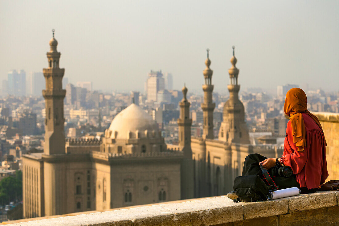 A young woman sits on a wall with a camera at the Great Mosque of Muhammad Ali Pasha looking out at the Mosque-Madrassa of Sultan Hassan and cityscape of Cairo,Cairo,Egypt