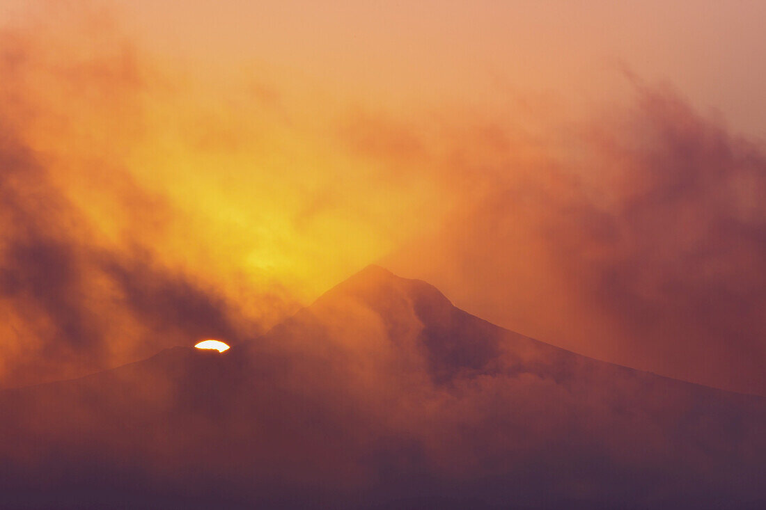 Dramatisch leuchtender Himmel in Gelb und Rosa mit Wolken, die die Silhouette des Mount Hood und seinen Gipfel verdecken, während die Sonne gerade noch hinter dem Berg sichtbar ist, Oregon, Vereinigte Staaten von Amerika