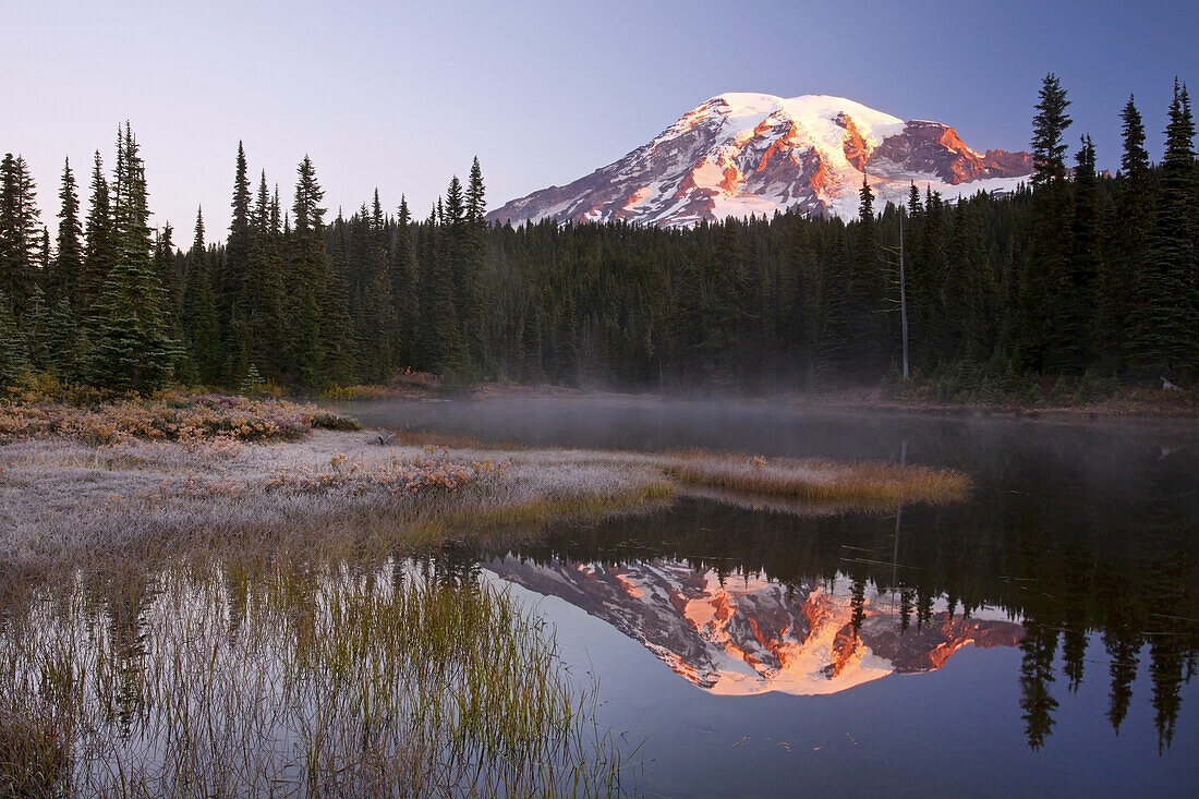 Mount Rainier at sunrise,with sunlight glowing on the snow-capped peak and the mirror image reflected in the tranquil water of Reflection Lake below,frosty grass on the shore and mist rising up from the water,Washington,United States of America