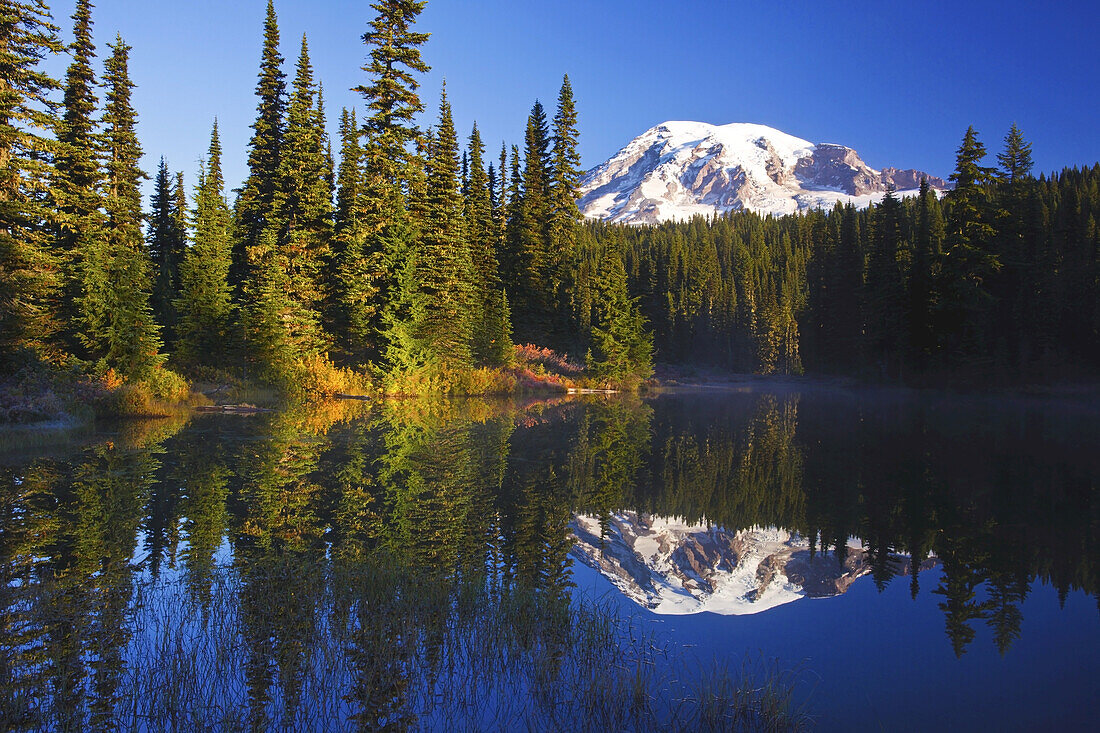 Mount Rainier bei Sonnenaufgang, mit Sonnenlicht, das den schneebedeckten Gipfel beleuchtet, und dem Spiegelbild, das sich im ruhigen Wasser des darunter liegenden Reflection Lake widerspiegelt, Washington, Vereinigte Staaten von Amerika