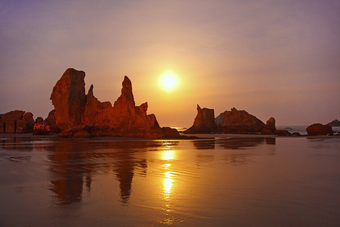 Zerklüftete Felsformationen entlang der Küste mit einer hellen Sonne, die bei Sonnenaufgang am Himmel leuchtet und sich auf dem nassen Sand in der Bandon State Natural Area an der Küste von Oregon, Bandon, Oregon, Vereinigte Staaten von Amerika, spiegelt