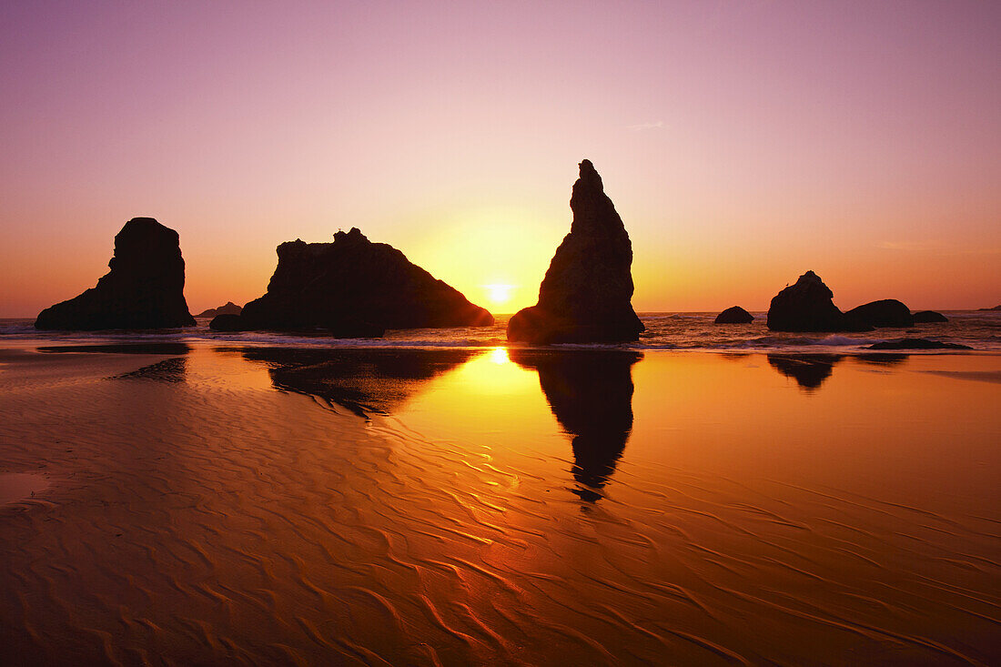 Rugged rock formations along the shoreline with a bright sun glowing in the sky at sunrise and reflecting on the wet sand at Bandon State Natural Area on the Oregon coast,Bandon,Oregon,United States of America