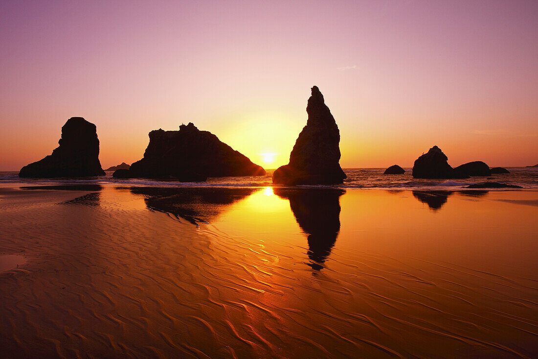 Zerklüftete Felsformationen entlang der Küste mit einer hellen Sonne, die bei Sonnenaufgang am Himmel leuchtet und sich auf dem nassen Sand in der Bandon State Natural Area an der Küste von Oregon, Bandon, Oregon, Vereinigte Staaten von Amerika spiegelt