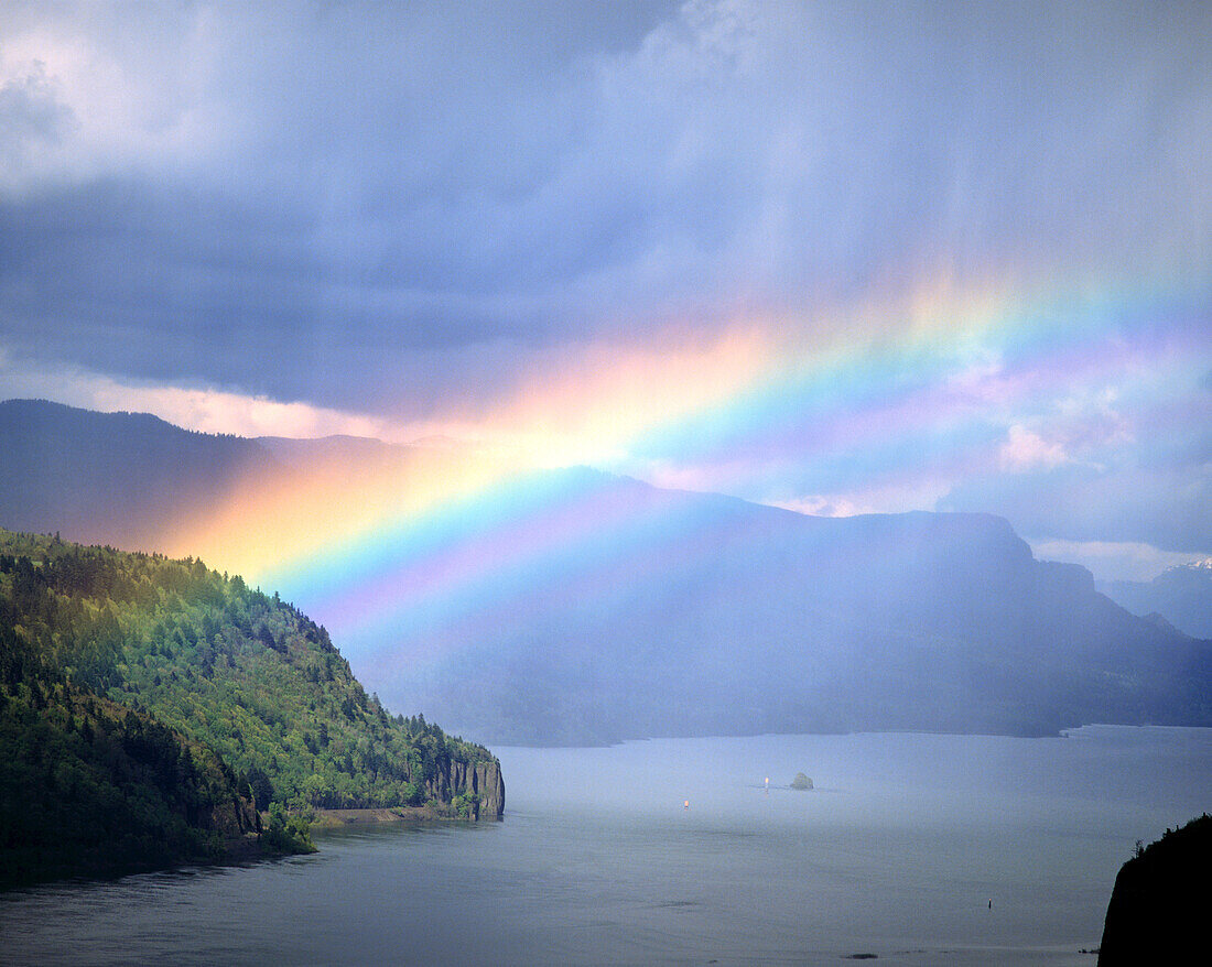 A bright rainbow shining through the rain clouds and rain over the Columbia River and shoreline in the Columbia River Gorge,Oregon,United States of America