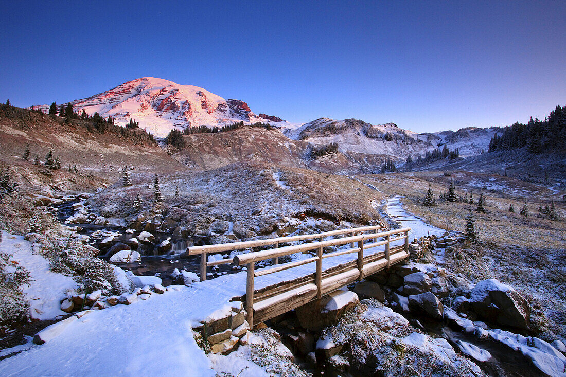 Brücke und Weg durch die Cascade Mountains im Winter, mit schneebedecktem Mount Rainier und Edith Creek im Paradise Park, Mount Rainier National Park, Washington, Vereinigte Staaten von Amerika