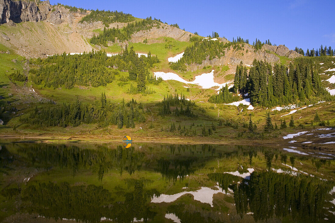 A tent set up on the water's edge of a tranquil Tipsoo Lake in the mountains with traces of snow on the slopes,Mount Rainier National Park,Washington,United States of America